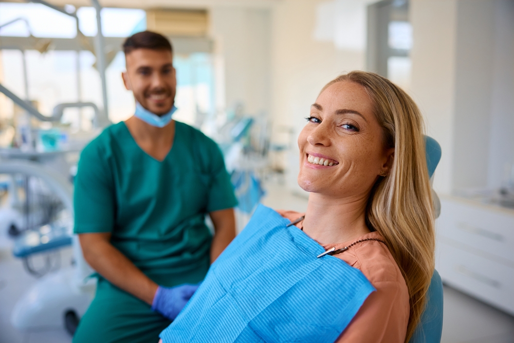 A woman with long blonde hair smiles in a dental chair, wearing a blue bib. Behind her, a male dentist in green scrubs and gloves smiles in a bright, modern dental clinic.